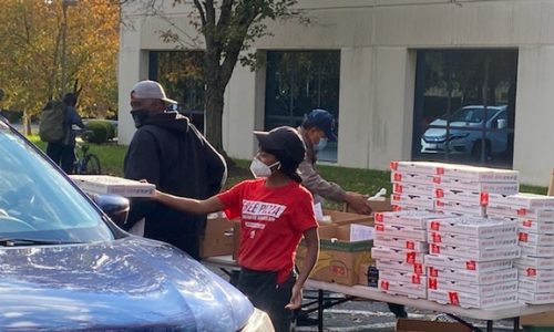 Volunteers distributing pizza boxes to cars at a community drive-through event.